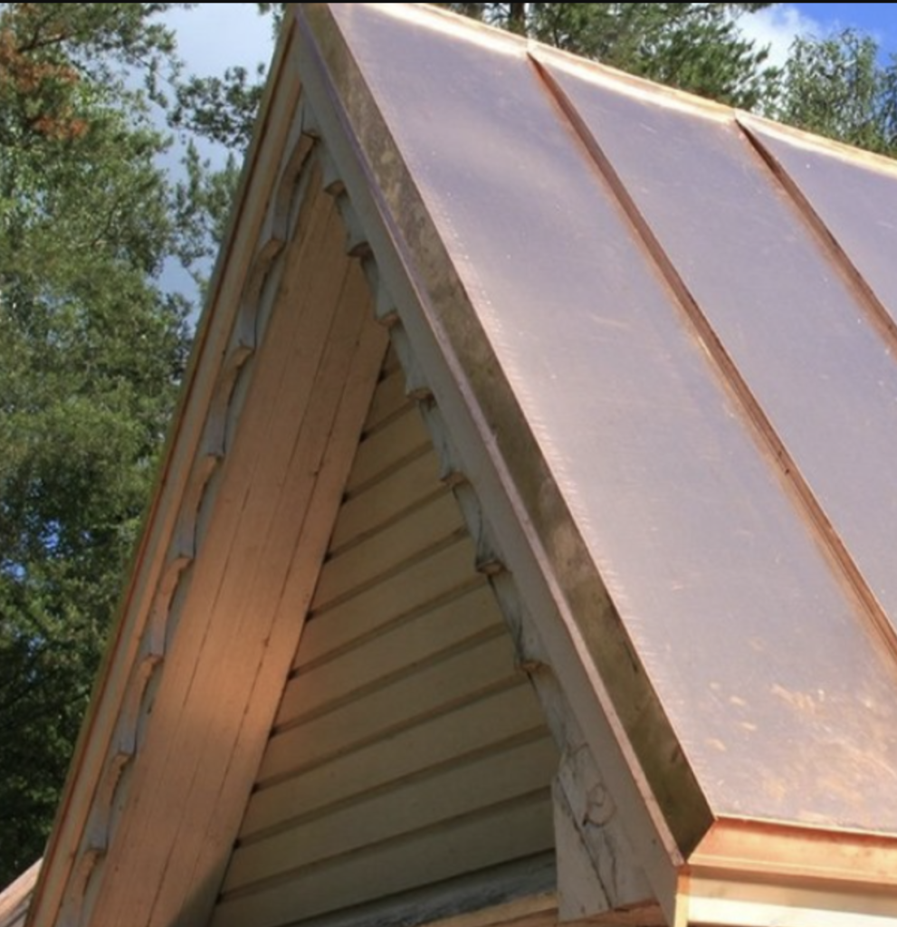 Screenshot 2024 05 21 at 2.45.52 PM 1 Close-up of a copper-colored standing seam metal roof on a residential structure, showing vertical seams and detailed roof edging against a wooded background.