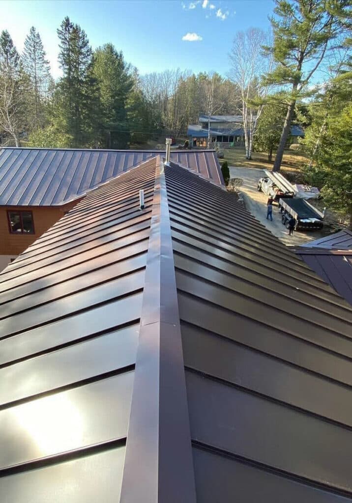 View from a roof ridge looking down at metallic panels, with trees, a road, a house, and a parked truck in the background.