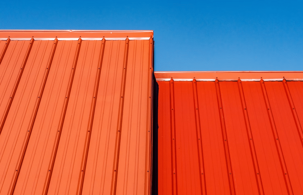 Two red metal rooftops, showcasing a striking metal roofing comparison—one lighter, one darker—intersect under a clear blue sky.