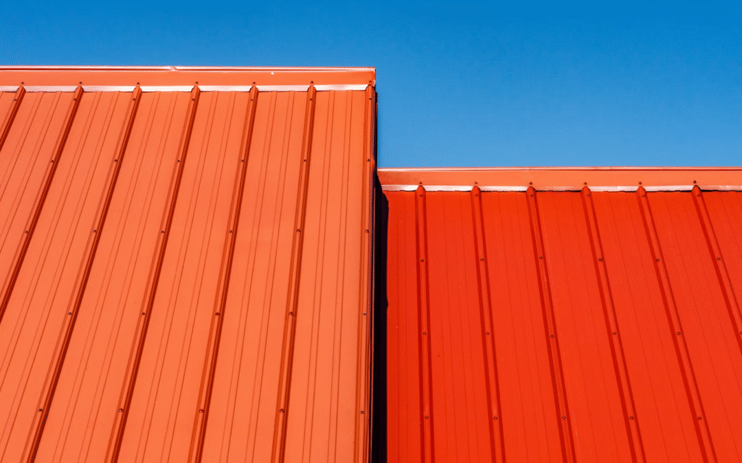 Two red metal rooftops, showcasing a striking metal roofing comparison—one lighter, one darker—intersect under a clear blue sky.
