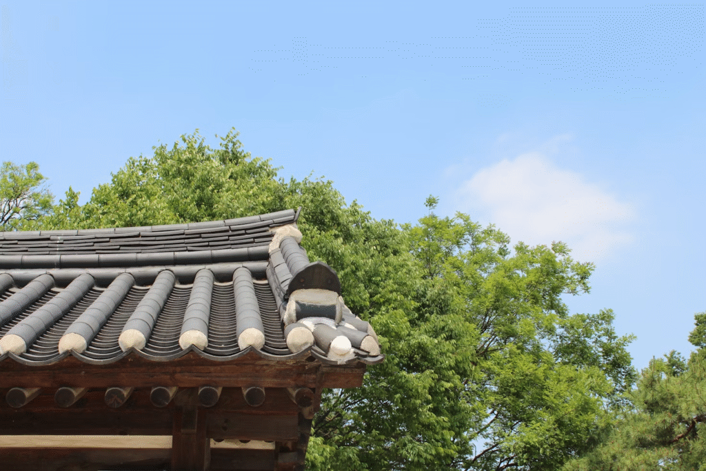 A traditional Korean tiled roof and wooden structure stands before green trees under a clear blue sky, offering a striking roofing comparison to modern metal roofing options.