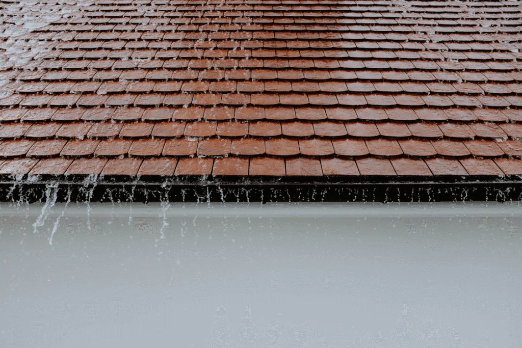 Water cascades off the edge of a sloped brown traditional roofing during rainfall, with streams visible against a white wall below.