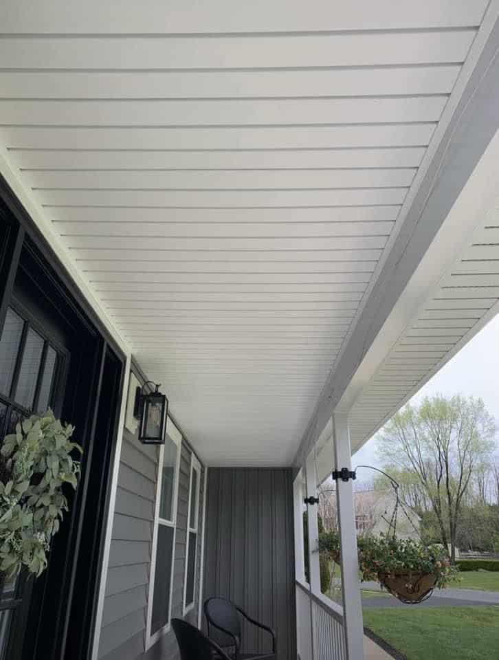 A covered front porch with white ceiling panels, a black chair, gray siding, a hanging planter, lantern light, and a view of trees and lawn outside.