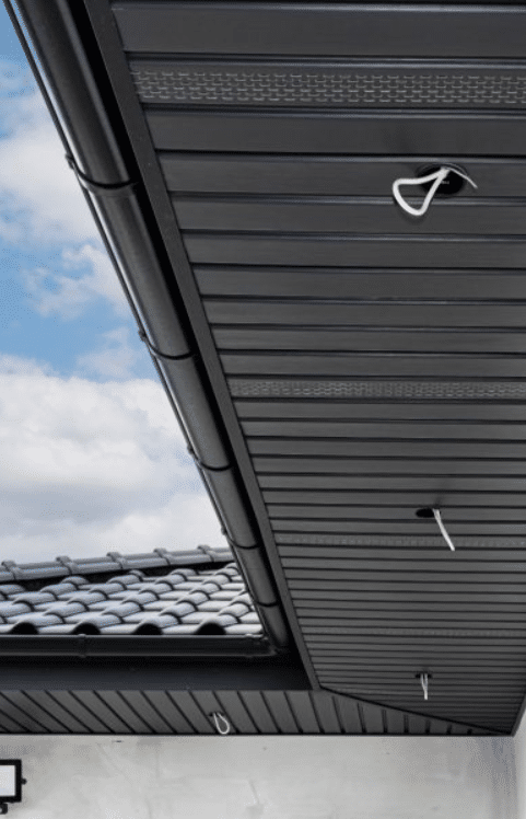 Black metal roof soffit with hooks installed, set against a partially cloudy blue sky and a tiled roof section in the background.