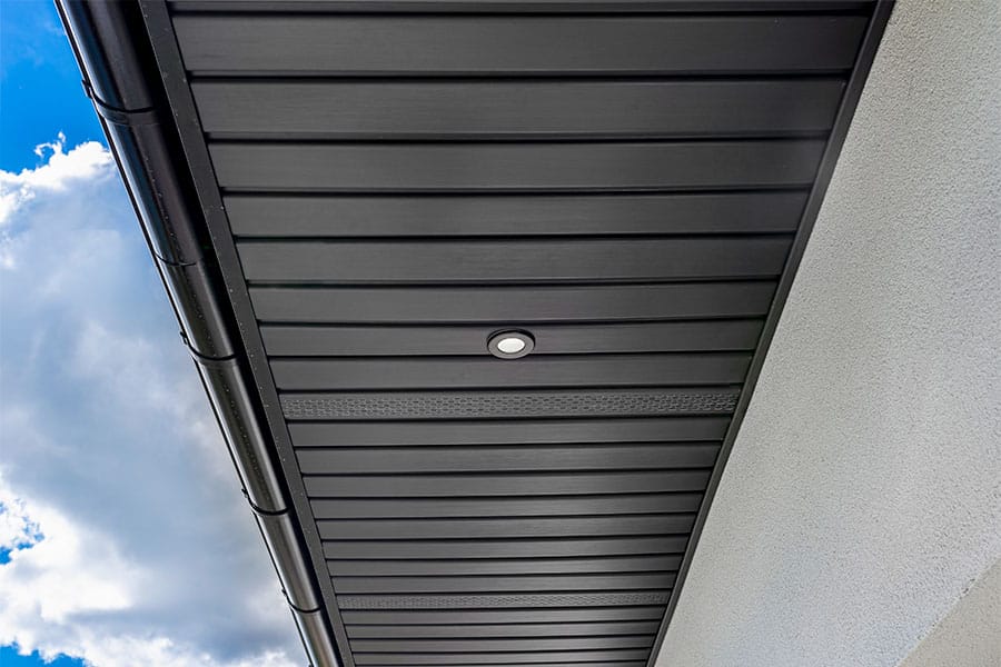Close up view of a modern house eave with black metal soffit panels, circular recessed light, white exterior wall, and partly cloudy sky in the background.