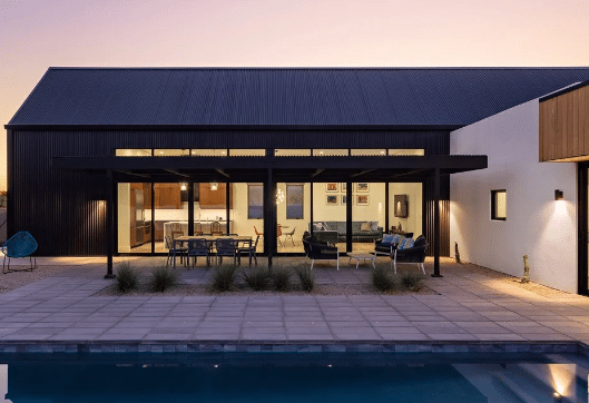 Modern house with large glass doors, outdoor seating under a pergola, and a swimming pool in the foreground at sunset—designed to withstand heavy snow like homes in The Rockies.