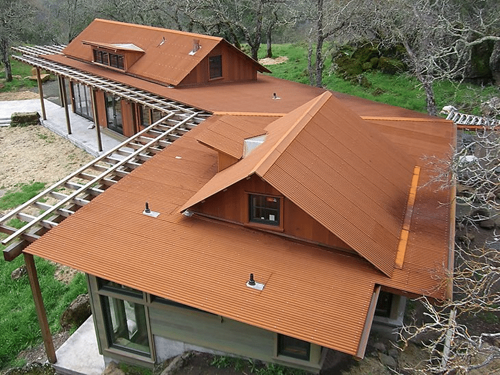 A modern house with rust colored mechanical seam metal roofing, large windows, and a wooden pergola, surrounded by grass and leafless trees—perfectly suited for heavy snow in The Rockies.