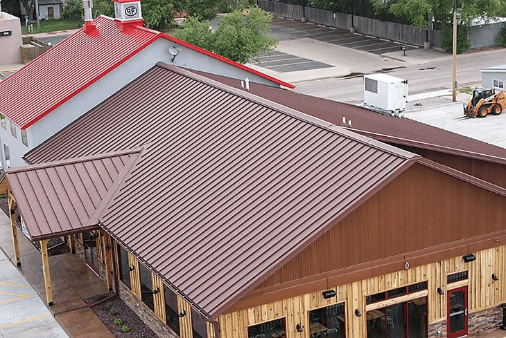 A building with a brown mechanical seam metal roof and wooden exterior sits next to another structure with a gray wall and red metal roof in a paved area, designed to withstand heavy snow common in the Rockies, with vehicles and trees nearby. A building with a brown mechanical seam metal roof and wooden exterior sits next to another structure with a gray wall and red metal roof in a paved area, designed to withstand heavy snow common in the Rockies, with vehicles and trees nearby.